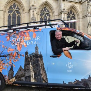 Dean Stephen sitting in the drivers seat of the e-van outside Gloucester Cathedral. The van is decorated with a picture of the Cathedral and autumn leaves against a vibrant blue sky