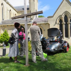 Reader Alexandra Dyer looks at a motorbike and sidecar with two friends, a cross is in the foreground and they are standing outside a church