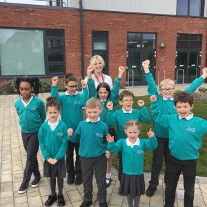 A group of children with their arms raised in celebration, wearing the teal green uniform of Clearwater school. A blonde woman's head is just visible in the back of the picture - this is the head, Kate Moss.