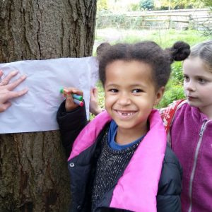 A girl smiles to the camera while she uses a crayon to take a rubbing from a tree