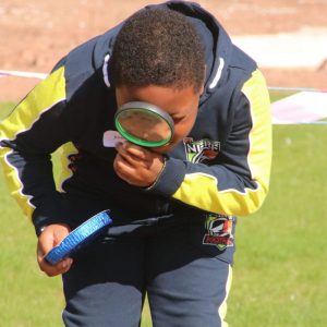 A boy wearing sports clothes bends towards the ground with a magifying glass to his eye